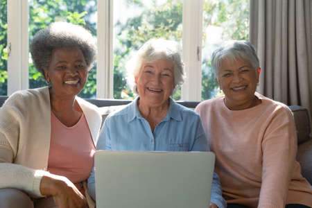 Three happy diverse senior woman sitting on sofa and using laptop. retirement lifestyle relaxing at home with technology.の写真素材