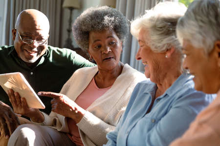 Three happy diverse senior woman and african american male friend sitting on sofa and using tablet. retirement lifestyle relaxing at home with technology.の写真素材