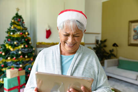 Happy mixed race senior woman in santa hat using tablet at christmas time. christmas, festivity and communicationの写真素材