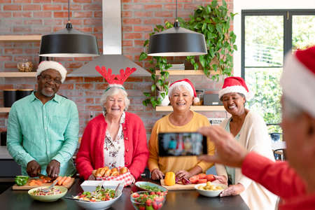 Group of happy diverse senior male and female friends in christmas hats taking selfie in kitchen. christmas festivities, celebrating at home with friends.の写真素材