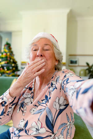 Happy caucasian senior woman wearing santa hat having video call, sending kisses at christmas time. christmas, festivity and communication technology.の写真素材