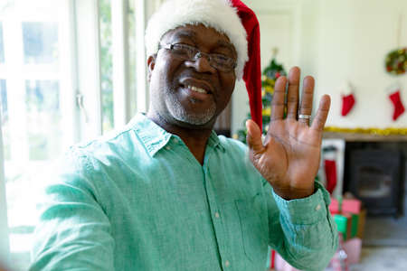 Happy african american senior man wearing santa has having video call at christmas time. christmas, festivity and communication technology.の写真素材