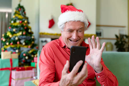 Happy caucasian senior man wearing santa hat having video call on smartphone at christmas time. christmas, festivity and communication technology.の写真素材