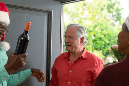 Happy caucasian senior man with wine coming to visit diverse friends at christmas time. christmas festivities, celebrating at home with friends.の写真素材
