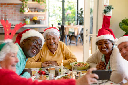 Diverse group of happy senior friends in holiday hats celebrating christmas together, taking selfie. christmas festivities, celebrating at home with friends.の写真素材
