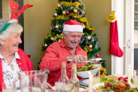 Happy caucasian senior couple in holiday hats sitting at christmas table. christmas festivities and traditions, celebrating at home.の写真素材