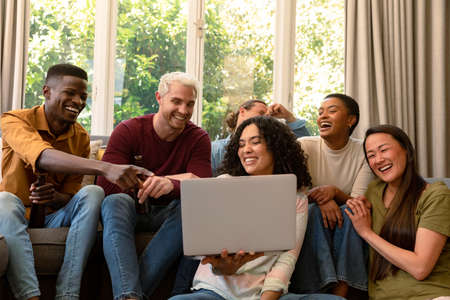 Group of happy diverse female and male friends drinking beer together and using laptop. socialising with friends at home.の写真素材