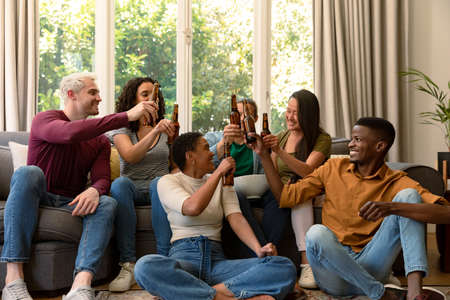 Group of happy diverse female and male friends watching tv and drinking beer together at home. socialising with friends at home.の写真素材