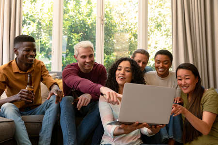 Group of happy diverse female and male friends drinking beer together and using laptop. socialising with friends at home.の写真素材