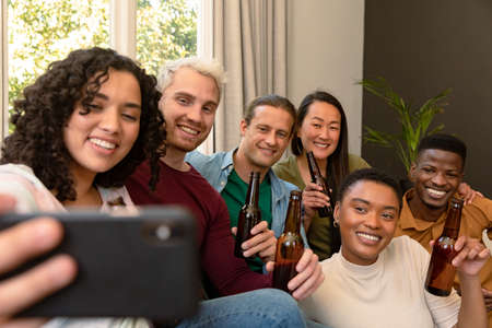 Group of happy diverse female and male friends drinking beer together and taking selfie. socialising with friends at home.の写真素材