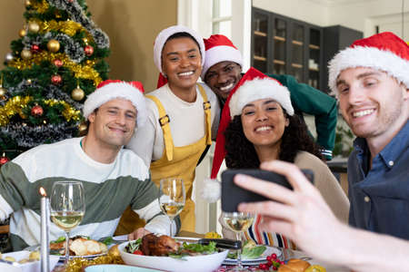 Group of happy diverse female and male friends in santa hats , celebrating christmas, taking selfie. christmas festivities, celebrating at home with friends.の写真素材