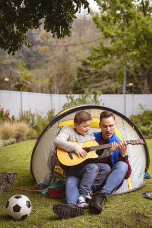 Caucasian father and son playing guitar together while sitting in a tent in the garden. fatherhood...