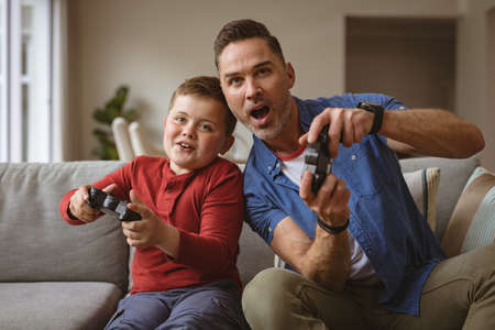Caucasian father and son playing video games together sitting on the couch at home. fatherhood and...