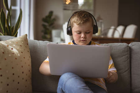 Caucasian boy wearing headphones using laptop sitting on the couch at home. home schooling and education conceptの写真素材