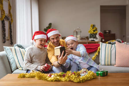 Caucasian father and two sons opening gift box during video call on digital tablet during christmas. social distancing during covid 19 pandemic at christmas timeの写真素材