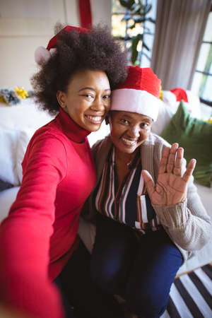 Smiling african american adult daughter and senior mother in santa hats taking christmas selfie. christmas, festivity and communication technology at home.の写真素材