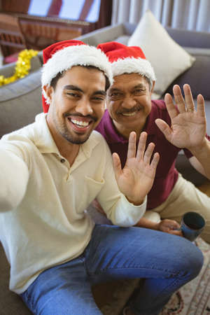 Portrait of happy biracial adult son and senior father in santa hats making christmas video call. christmas, festivity and communication technology.の写真素材