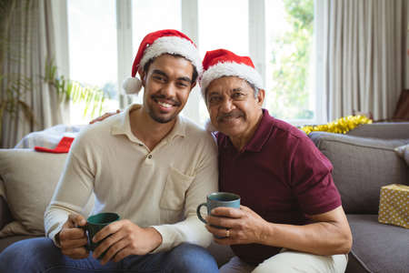 Portrait of happy biracial adult son and senior father in santa hats making christmas video call....