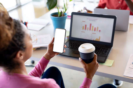 Businesswoman holding coffee cup while looking at smartphone with copy space in office. business,...