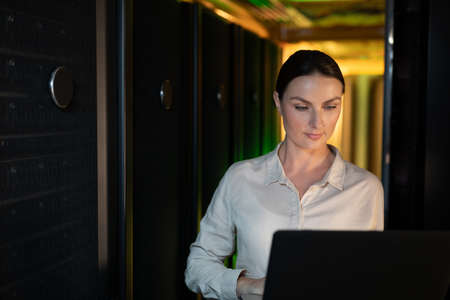 Caucasian female engineer using laptop in computer server room. database server management and maintenance conceptの写真素材