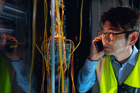 Asian male engineer talking on smartphone while inspecting in computer server room. database server...