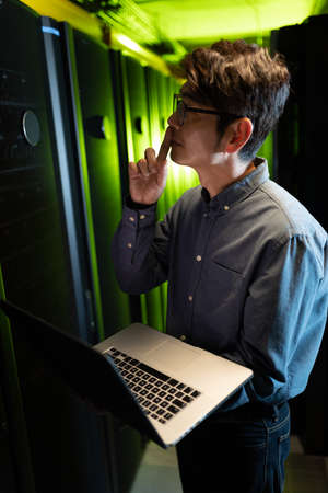 Thoughtful asian male engineer with laptop inspecting in computer server room. database server management and maintenance conceptの写真素材