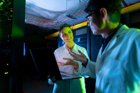 Diverse male and female engineers wearing aprons discussing over laptop in computer server room....