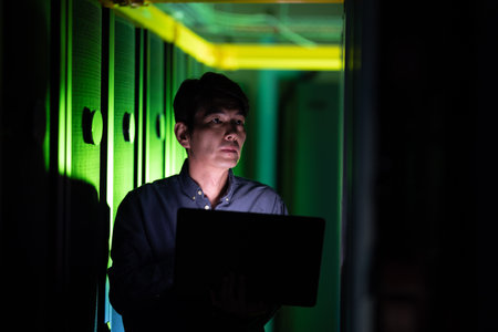Asian male engineer using laptop while inspecting in computer server room. database server...