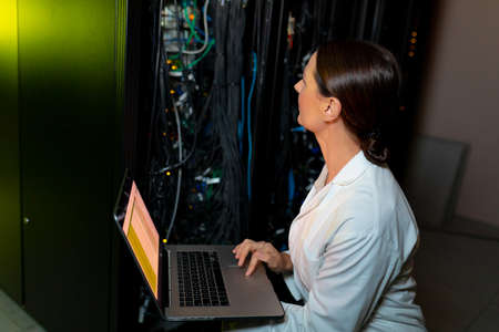 Caucasian female engineer wearing an apron using laptop while inspecting in computer server room. database server management and maintenance conceptの写真素材