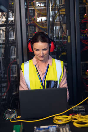 Caucasian female engineer wearing ear plugs using a laptop in computer server room. database server management and maintenance conceptの写真素材