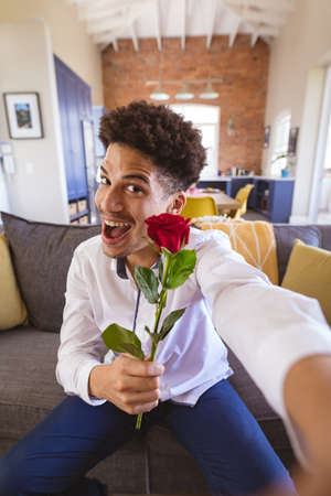 Portrait of biracial young man with rose enjoying virtual date at home. lifestyle and virtuality.の写真素材
