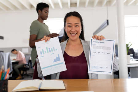 Portrait of smiling asian creative businesswoman showing charts while sitting at desk in office....