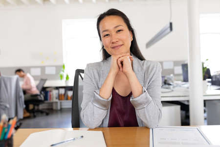 Portrait of confident asian creative businesswoman sitting with hands on chin at desk in office....