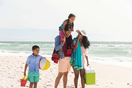 Happy biracial man carrying daughter on shoulders while walking with woman and son at beach. family, lifestyle and weekend.の写真素材