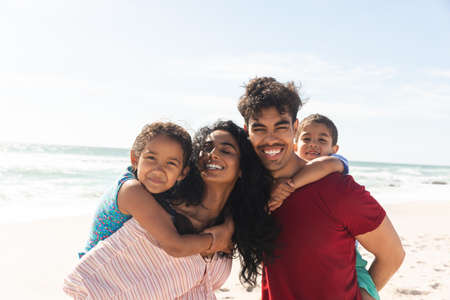 Happy biracial father and mother giving piggyback rides to kids at beach against sky with copy...