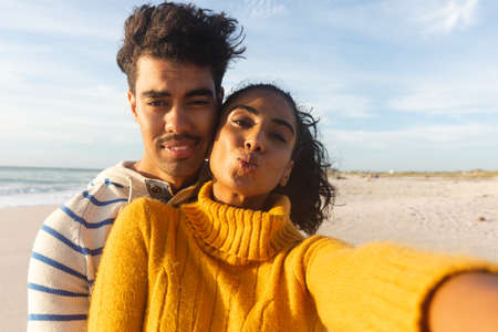 Portrait of young biracial woman puckering while taking selfie with boyfriend at beach on sunny day. lifestyle, love and weekend.の写真素材