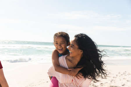 Happy biracial woman giving piggyback ride to daughter at beach against sky with copy space. family,...
