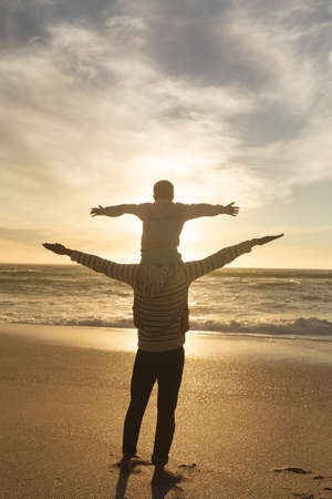 Rear view of biracial man carrying son on shoulders with arms outstretched at beach during sunset. family, lifestyle and weekend.の写真素材