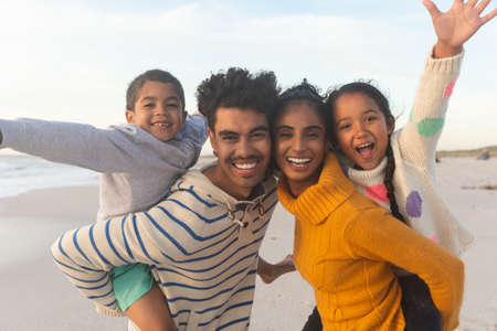 Portrait of happy biracial father and mother giving piggyback rides to kids at beach during sunset. family, lifestyle and weekend.の写真素材