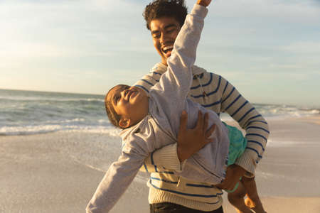 Cheerful biracial man carrying daughter pretending to fly at beach against sky on sunny day. family,...