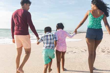 Rear view of biracial parents holding hands with children walking on sand at beach during sunny day. family, lifestyle and weekend.の写真素材