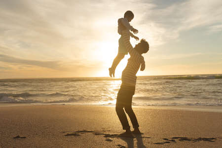 Side view of playful biracial father holding son aloft on shore at beach against sky during sunset. family, lifestyle and weekend.の写真素材