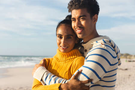 Smiling biracial man embracing girlfriend while looking away at beach on sunny day. lifestyle, love and weekend.の写真素材