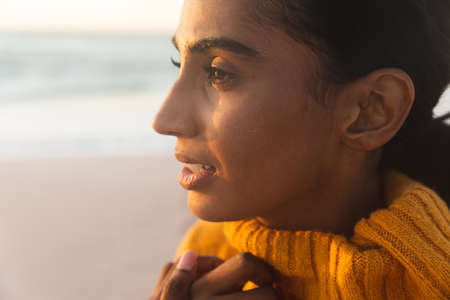 Close-up of young thoughtful biracial woman in sweater looking away at beach during sunset. lifestyle and weekend.の写真素材