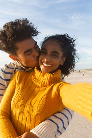 Portrait of smiling biracial woman taking selfie of boyfriend hugging and kissing on cheek at beach. lifestyle, love and weekend.の写真素材