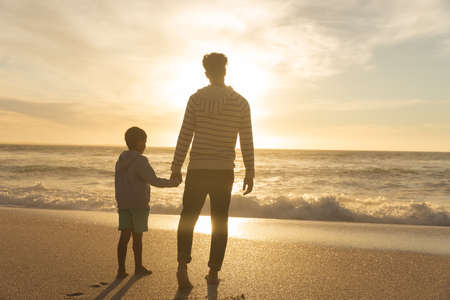 Full length rear view of biracial father and son holding hands while looking at sunset over sea. family, lifestyle and weekend.の写真素材