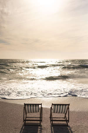 Two empty folding wooden chairs on shore in front of waves splashing in sea at beach against sky. nature and relaxation.の写真素材