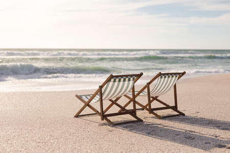 Two empty folding wooden chairs on shore at beach in front of waves splashing in sea against sky. nature and relaxation.の写真素材