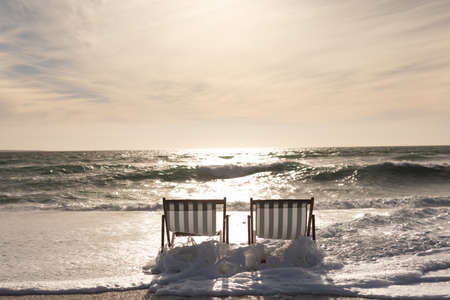 Wave splashing on empty two folding wooden chairs at beach against sky during sunset. nature and...