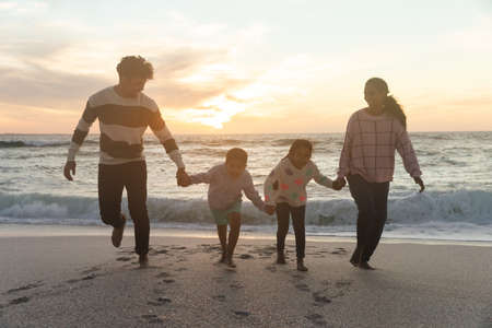Full length of happy multiracial family holding hands running at beach against sky during sunset. lifestyle and weekend.の写真素材